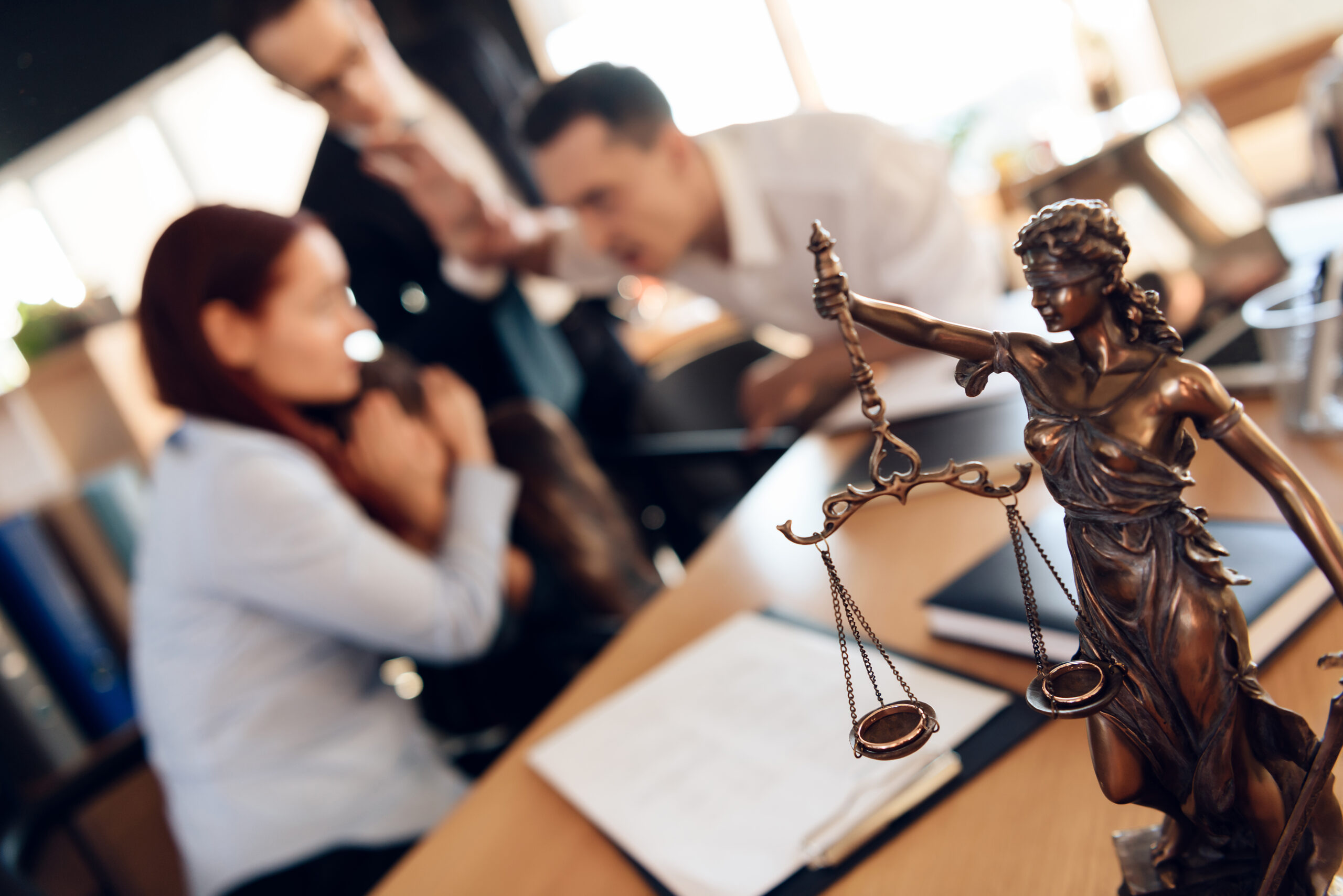 Bronze figurine of Themis holds wedding rings on scales of balance, being in foreground. Couple divorce. Adult couple gets divorced from attorney for divorce in office.